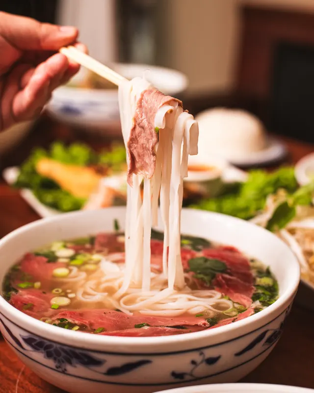 Chopsticks pulling rice noodles and rare beef out of a bowl of pho