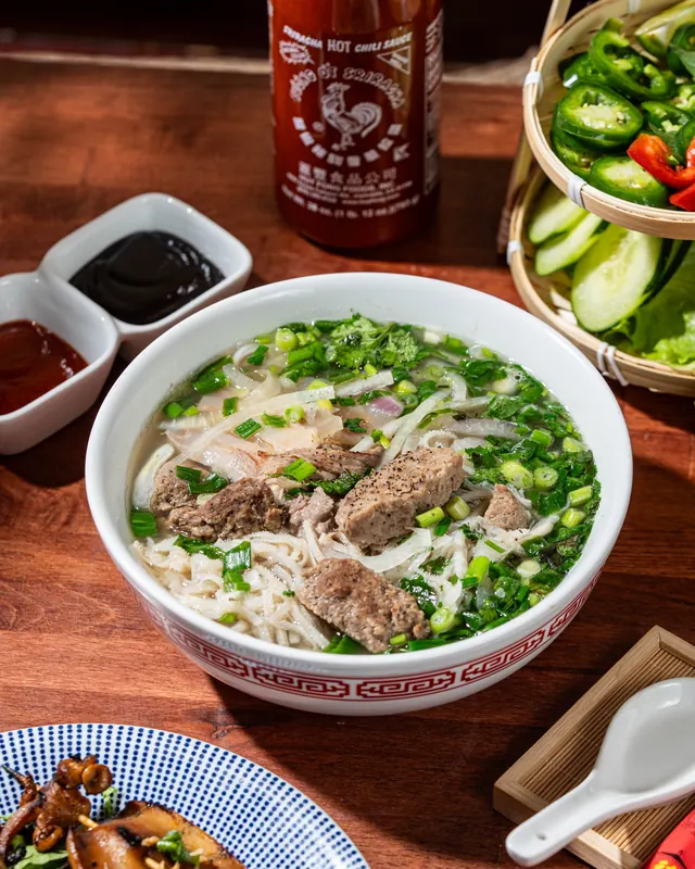 A bowl of pho with rare beef and meatballs, hoisin and sriracha on the side